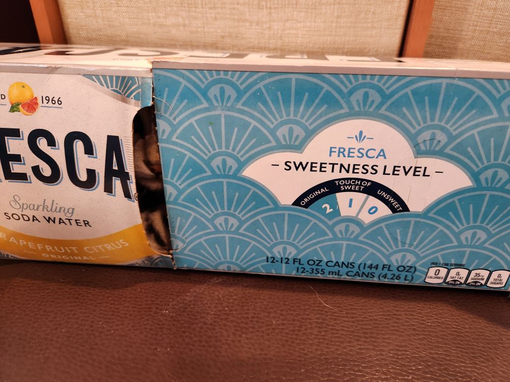 A fresca 12-pack box with a little bit if cat muzzle and eye seen through a gap.
