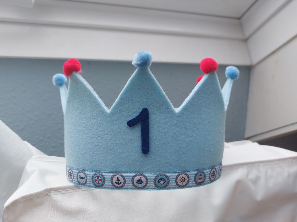 Hellblaue Geburtstagskrone aus Filz, auf den Spitzen blaue undrote Bömmelchen. Am unteren Rand eine Borte mit maritimen Motiven.