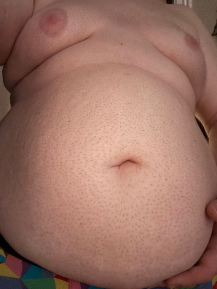 @bonniebilad’s soft body, with their gut protruding out. The camera is facing upwards at their looming, hungry mass. they are wearing rainbow underwear, largely hidden under their belly. Their breasts are up far away from the viewer, but clearly soft and pushed forward, their pink nipples puffy and soft. A roll of fat is squished between their gut and tits. Their hand holds the bottom of their distended, smooth gut, awaiting more weight to fill itThe lighting is somewhat dim but warm.