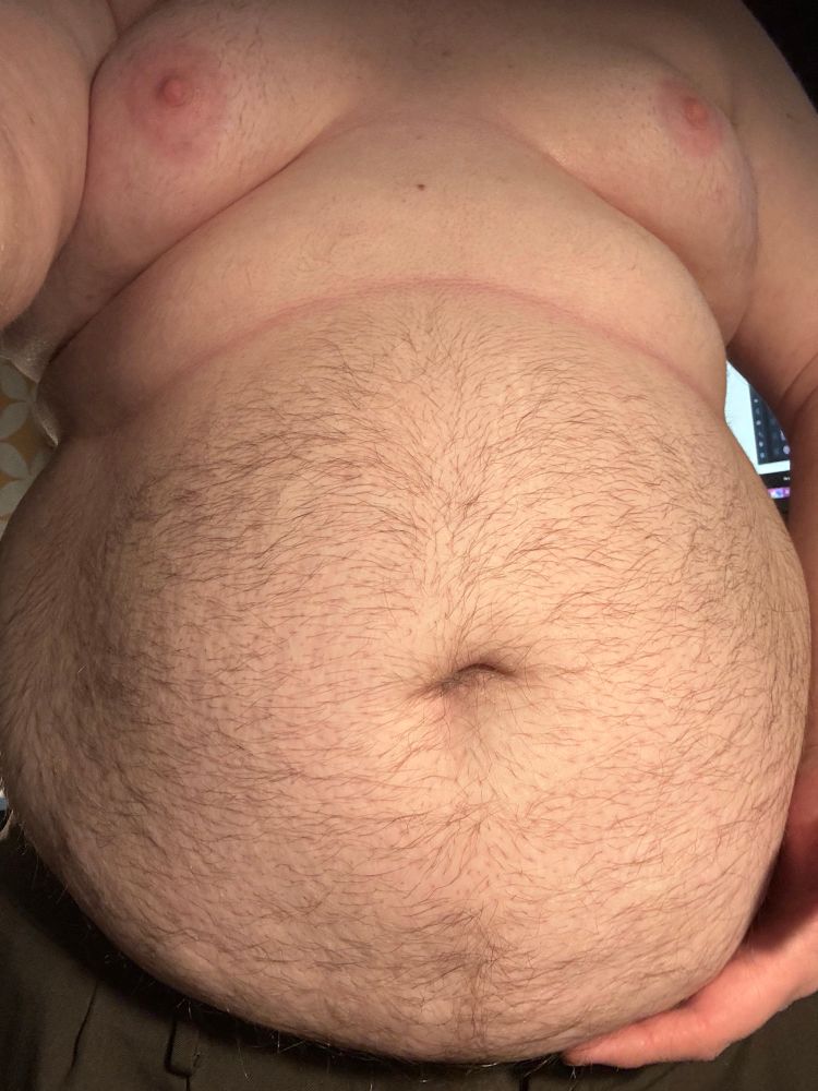 @bonniebilad’s hairy belly, illuminated with bright warm light. The camera is nearly straight-on, showing the dimpling at the bottom of their gut. A neat roll of fat with a crisp crease line is above their gut, oozing to the size, and their breasts are perked out with bright pink nipples. 
