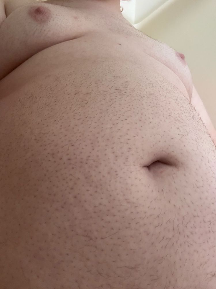 @bonniebilad’s soft and filled body, their breasts sticking out little as their  bloated, full gut sticks out far past them, with a roll of fat pulled taught between them. The lighting is at an angle and quite pale on their pale skin.