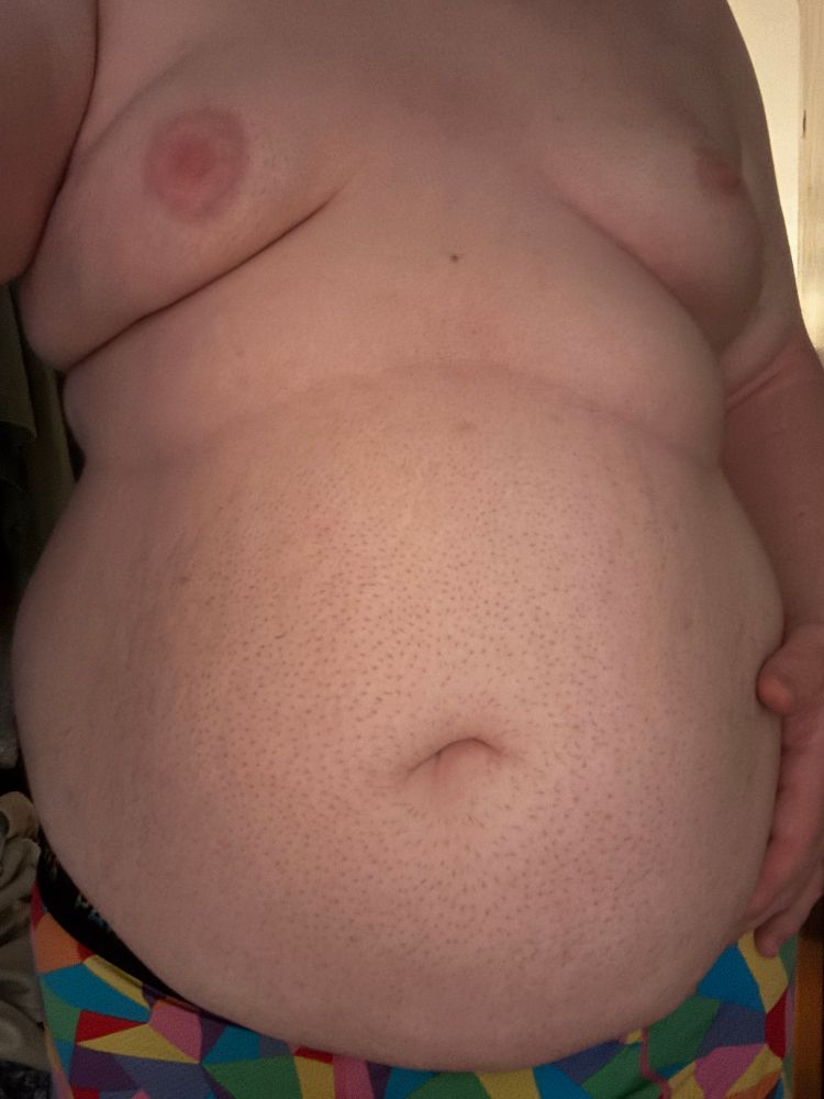 @bonniebilad’s soft body, with their gut protruding out. they are wearing rainbow underwear, largely hidden under their belly. Their breasts are soft and pushed forward, their pink nipples puffy and soft. A roll of fat is visible between their gut and tits. Their hand rests on their distended, smooth gut. The lighting is somewhat dim but warm.
