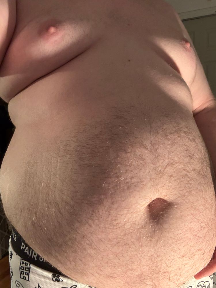 @bonniebilad’s heavy belly, hanging low off of their body, with warm lighting shining upwards at them. Their breasts jut out with broth pink nipples, and a roll of fat oozes out on top of their hungry gut.  Their belly button is shadowed, wide and deep. 