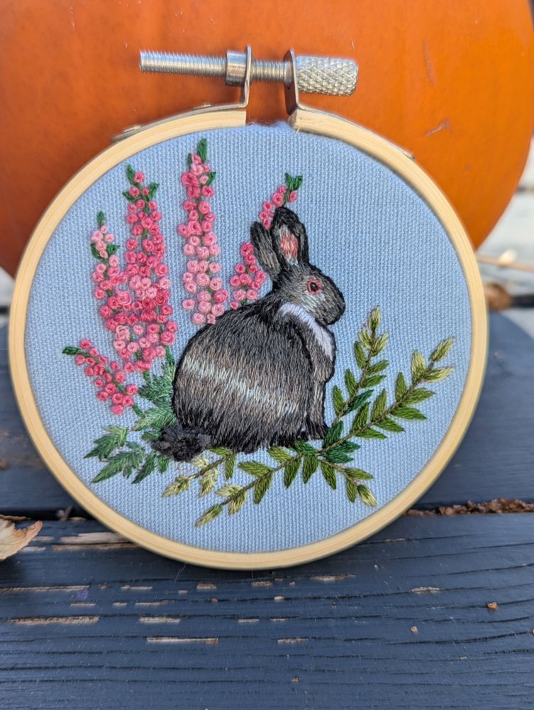A gray and black and white bunny stitched in long and short stitch on blue fabric. The bunny is surrounded by pink larkspur made of French knots and stem stitch, and shades of green foliage in satin stitch and stem stitch.