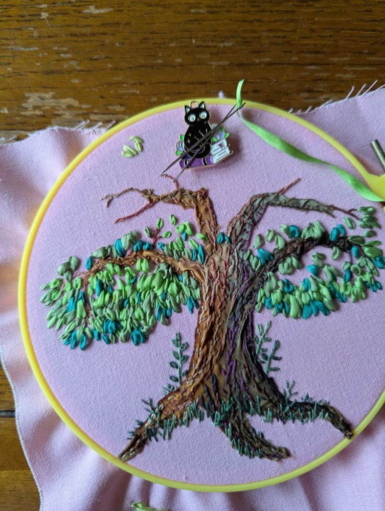 An embroidery hoop with pink fabric, upon which is stitched a tree trunk in ribbon with floss embellishments. Variegated green cotton floss is stitched around the roots in grass and little plants. Different shades of green silk ribbon have been stitched in the beginning of foliage