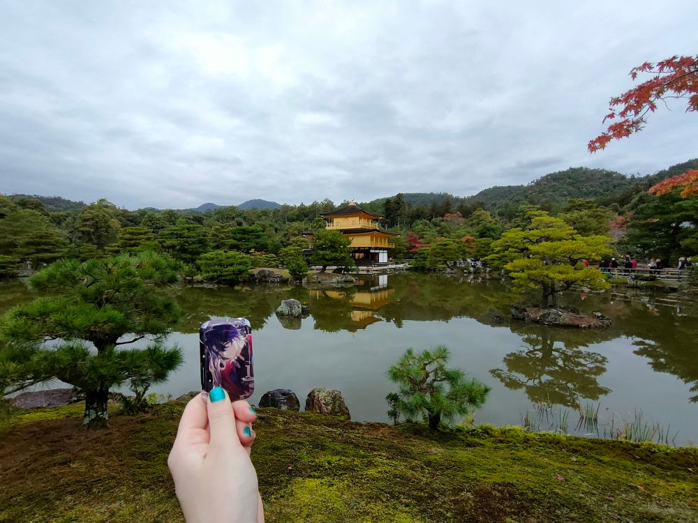 曇り空の金閣寺（水面にも反射している）と崩壊スターレイルのキャラクター、サンポのスクエア缶バッジを一緒に撮った写真。右側には紅葉が少しだけ顔をのぞかせている。