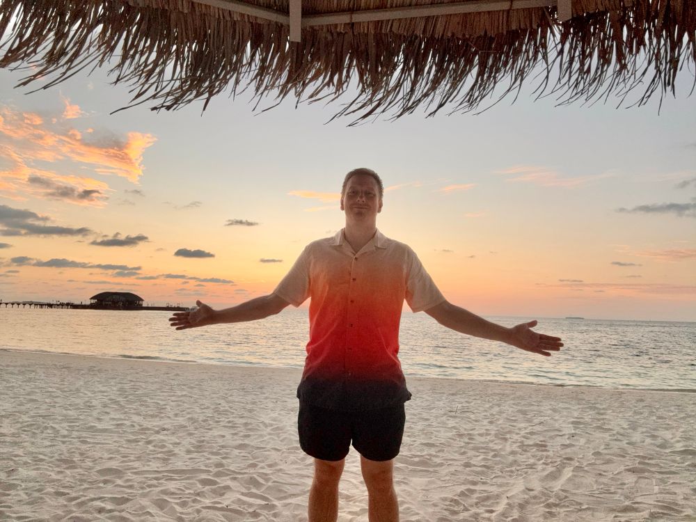 Dan is on a beach wearing a shirt that starts cream at the top, turning to orange in the middle and dark orange at the bottom, with black shorts underneath. He blends in with the sunset behind very well.