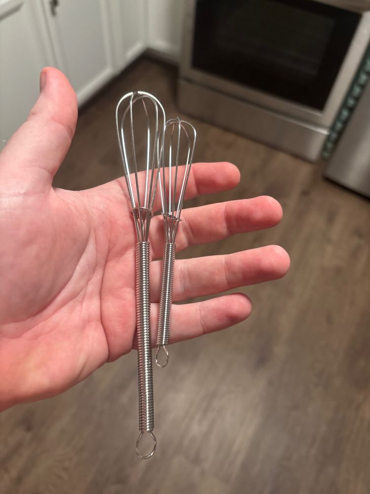 a man’s hand holding two tiny whisks, one smaller than the other