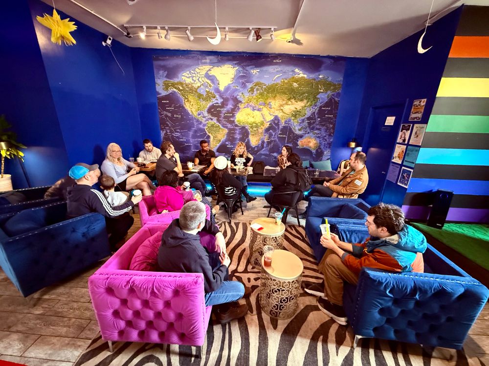 A vibrant room with blue walls featuring a large world map mural. A diverse group of people is sitting on colorful couches and chairs arranged around small tables. The seating includes bright pink, blue, and black sofas. The floor has a zebra-striped rug, and a multicolored wall is visible to the right. The atmosphere appears relaxed and social, with individuals engaged in conversation or activities.