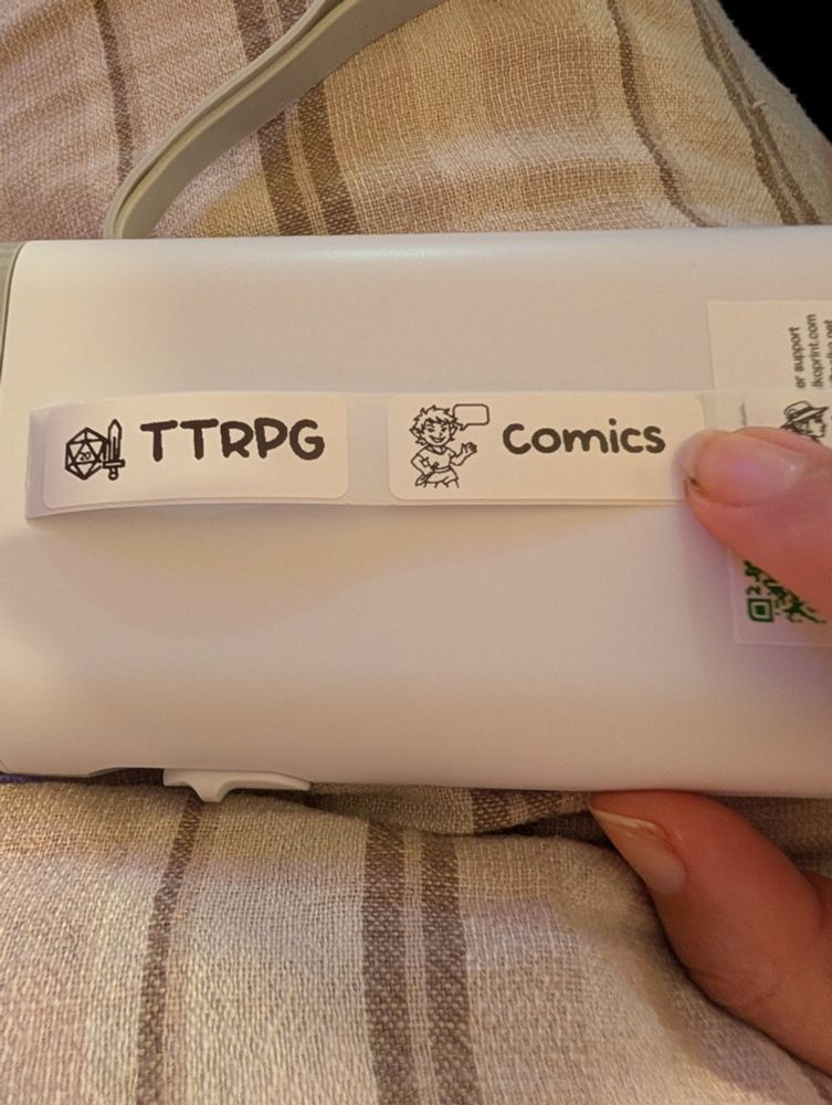 A close-up photo of a white handheld label printer resting on striped fabric. Two black-and-white printed labels are affixed to the printer. The first label on the left reads "TTRPG" with an icon of a sword and a 20-sided die. The second label reads "Comics" and features a cartoon-style character speaking in a word bubble.