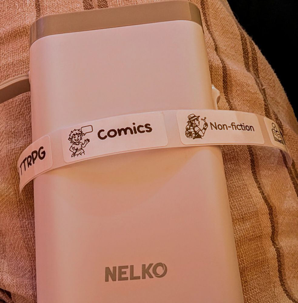 A wider shot of the same label printer, now displaying a full sequence of black-and-white labels wrapped around it. From left to right, the labels read: "TTRPG" with a sword and die icon, "Comics" with the speaking cartoon character, and "Non-fiction" with an icon of a person in explorer gear holding a pencil and scissors, suggesting science and crafting themes.