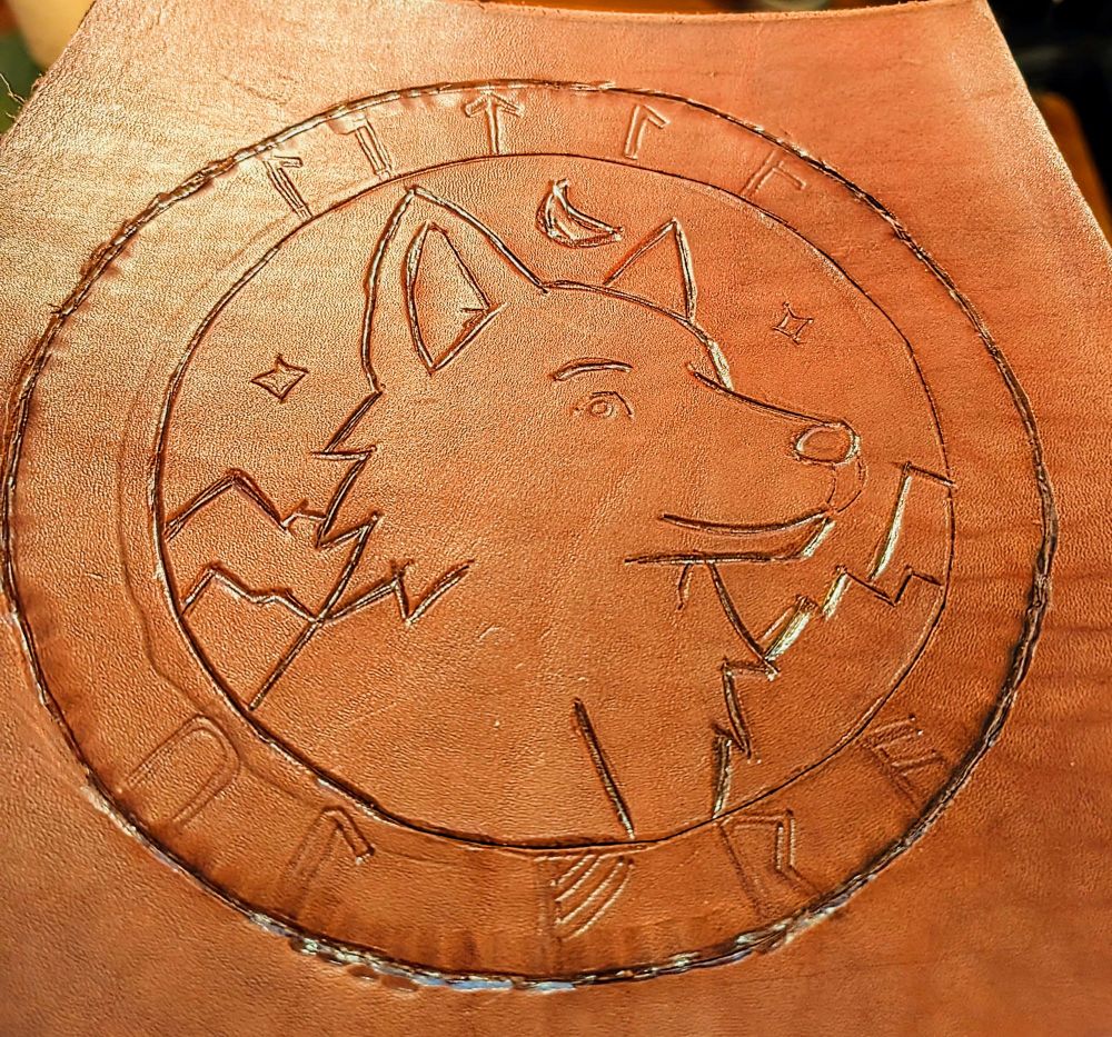 A piece of brown leather with a hand-tooled design featuring a stylized wolf head in profile, surrounded by a circular border. Inside the circle are mountain peaks, stars, and a crescent moon above the wolf’s head. The outer ring is engraved with Norse-style runes spaced evenly along the perimeter. The tooling is crisp and partially beveled, with visible knife and stamp marks indicating work in progress. The leather has a warm, reddish-brown tone and is photographed under warm lighting.