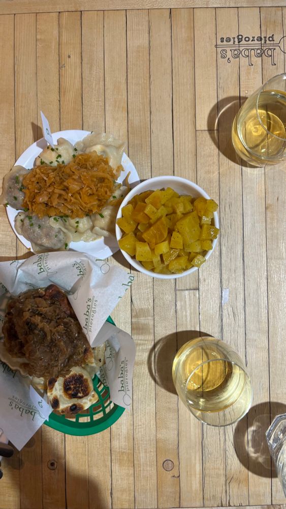 An overhead shot of pierogis, golden beet salad, and two glasses of white wine on a table made of pale strips of wood. The boiled pierogis have chives and sauerkraut on top; the pan fried pierogis have caramelized onions. The Baba’s Pierogis logo is visible on the corner of the table. 