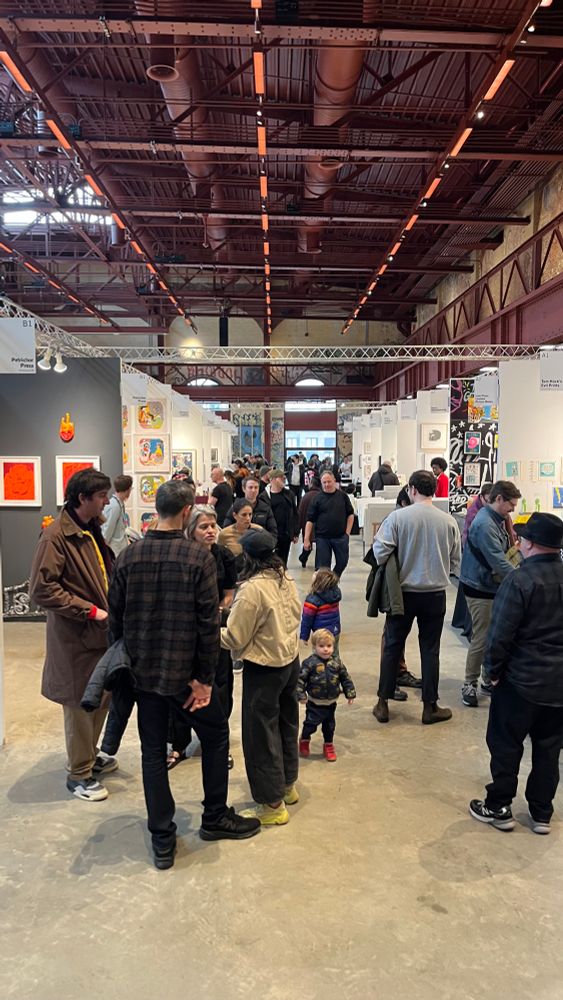 A shot of one of the aisles of booths at the Brooklyn Art Print Fair. It’s located in Powerhouse Arts, which is an old power plant covered in graffiti that they turned into an event/art space, so it’s very industrial-looking, with red metal beams, brick walls, and a concrete floor. Lots of people in the foreground of the photo and cool art hanging in the booth in the background.