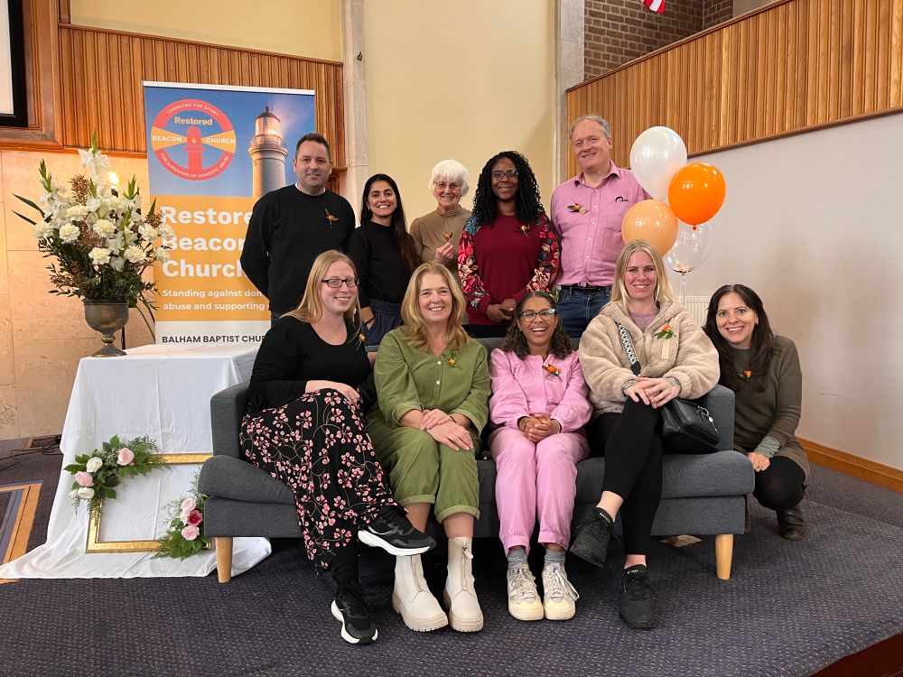 A group of 10 people in two rows, some seated on a grey sofa, others stood, next to orange, white and gold balloons and white floral displays, on a church stage. 