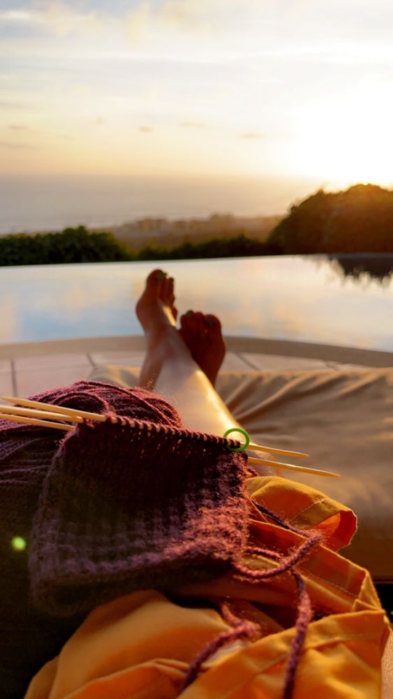 Image of Jen’s legs outstretched by the pool, with yarn and knitting materials in her lap and half of a glove worked up. The sun is setting, the image is calm and pleasant.