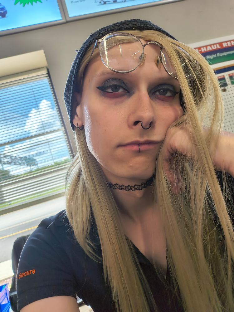Amy slumped at work
I have my glasses on my forehead and my cheek resting against my fist 
I have black top eyeshadow and red bottom, small wings 
black choker, black lip, nose and ear piercings 
hair all over the place, pinned by my fist 