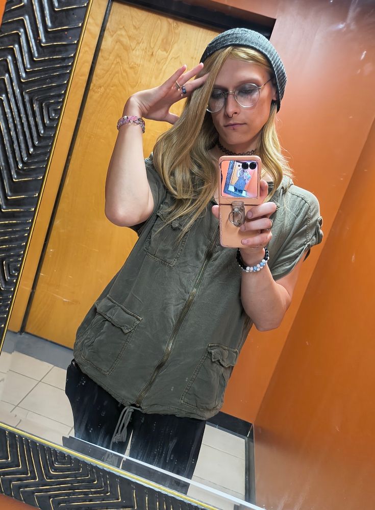 amaranth in the work bathroom mirror
she's wearing black ribbed leggings, a strange, baggy green top, grey hat, black facial jewelery
black eyeshadow on top, matching green on the bottom 
she has her hand flat up to her head, pointer finger resting on her forehead
