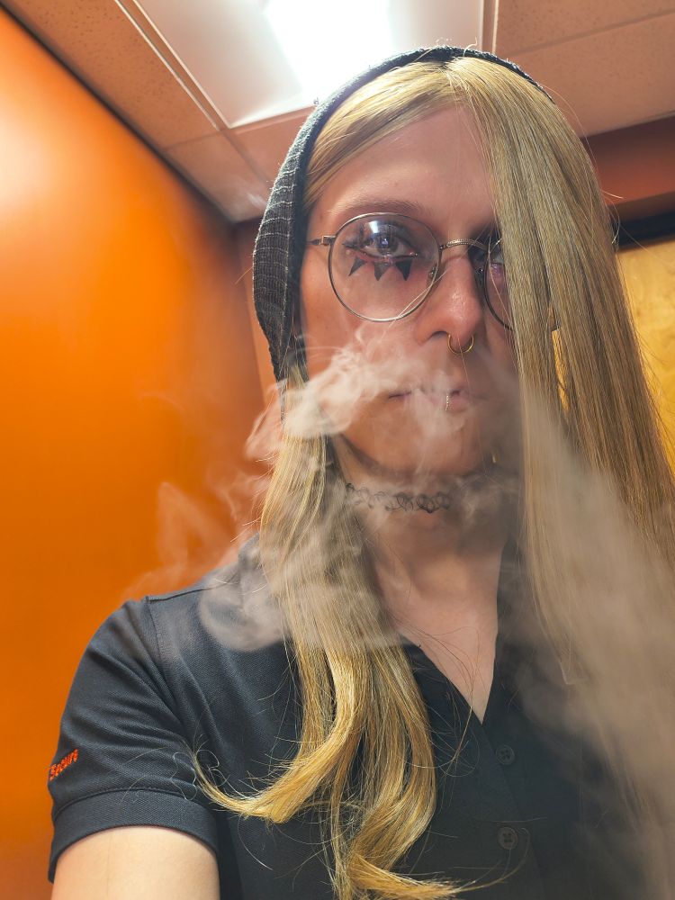 Amy in da work bathroom
she has a grey hat pulled over her ears, blonde curly ended hair 
she has three eyeliner spikes under her visible eye, the other is covered by hair
grey framed glasses, black choker, black work shirt 
