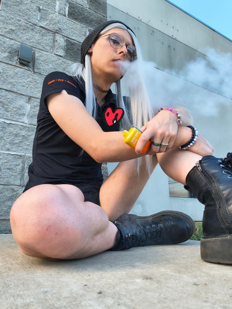 Amy sitting outside'a work 
she's wearing her black work shirt and black shorts and black boots, she has a yellow vape in her left hand, which is sitting on top of her right hand on her knee
she's got a black choker, jewelery and eyeshadow
she has rings on her middle and ring finger on her left hand, two bracelets on the right a pink bracelet on the left 
she hittin dat shit