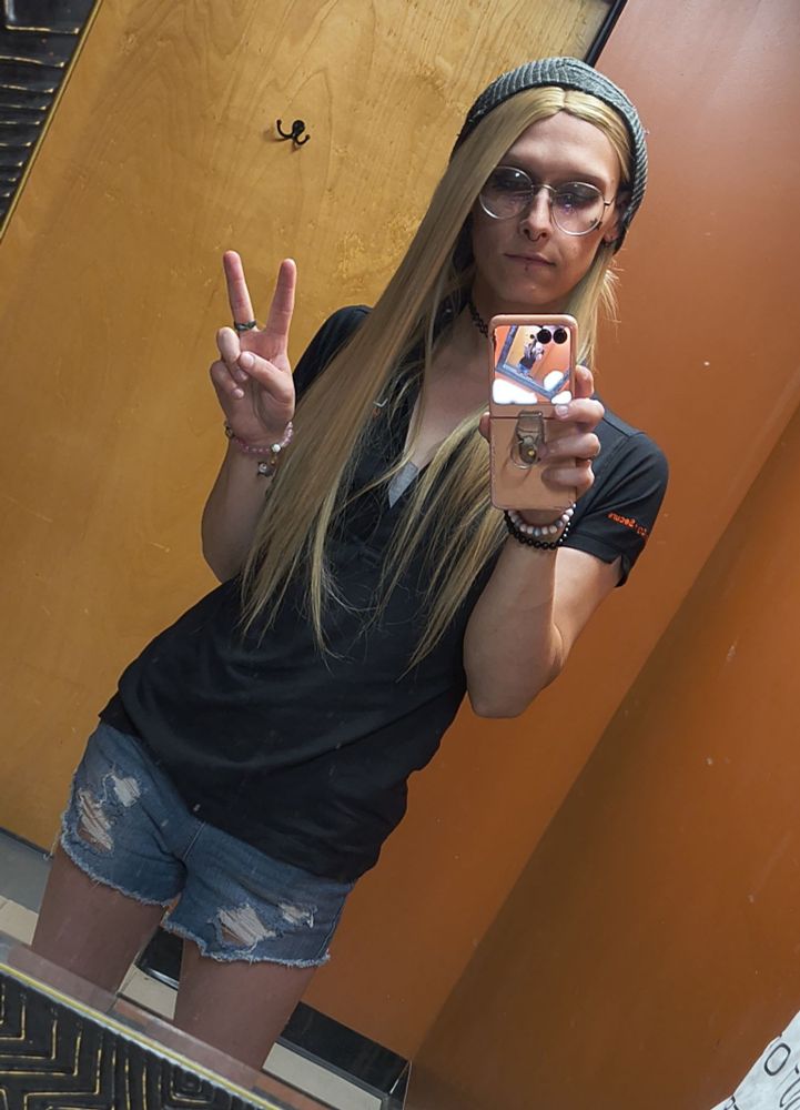 Amy in da work bathroom 
she's wearing torn up jean shorts, black work shirt, black hat 
she has her big round glasses on, a ring visible on her right middle finger, a pink bracelet on her right wrist and black and white bracelets on the left 
her phone case is pink with a ring hanging from the back of it in her left hand
holding up the peace sign 