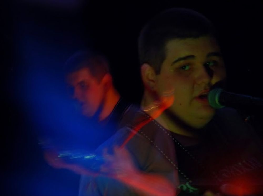 A performance image of Skeeball Pro. A person on a dark stage in a gray shirt and a guitar strap singing into a microphone. Another still of that person is superimposed on top with artful red and blue lighting. 