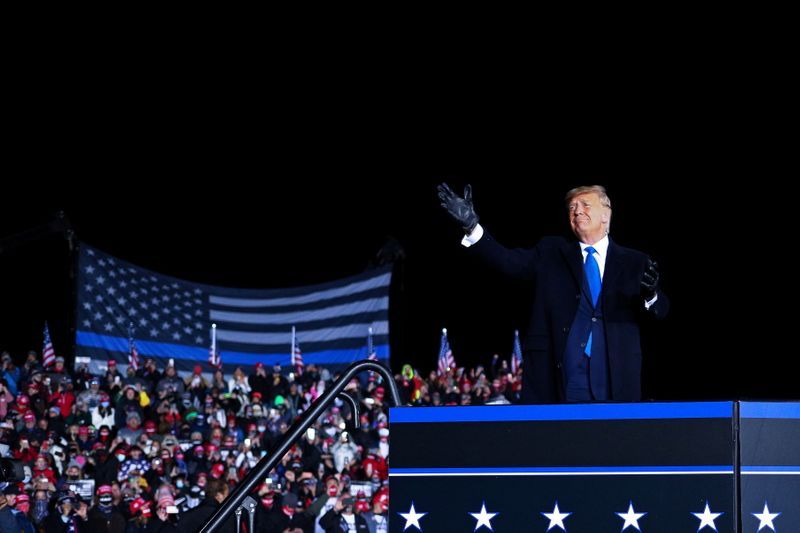 Trump wearing black gloves, black long coat, at a rally with a large "blue lives matter/thin blue line" flag