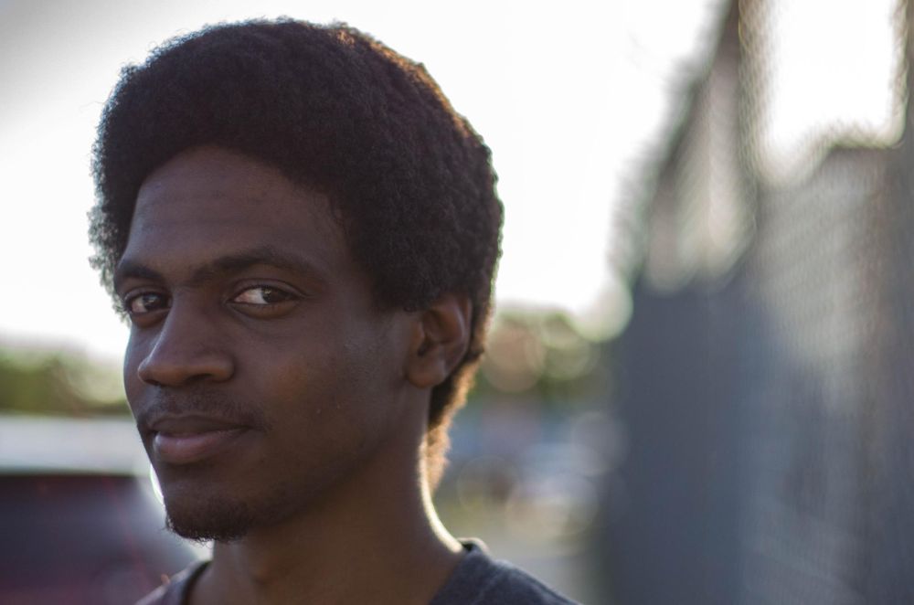 portrait, Jeremy Eugene; in the background, the fuzzed out barbed wire and chain link of the jail where we lost Sandra Bland; Jeremy is facing us but at an angle - his right cheek, hidden; he seems: confident, daring, fierce