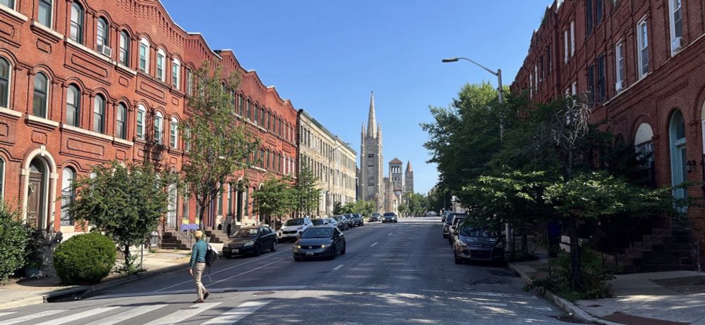 both sides of the street are lined with red brick rowhomes and many trees; in the distance, three church spires