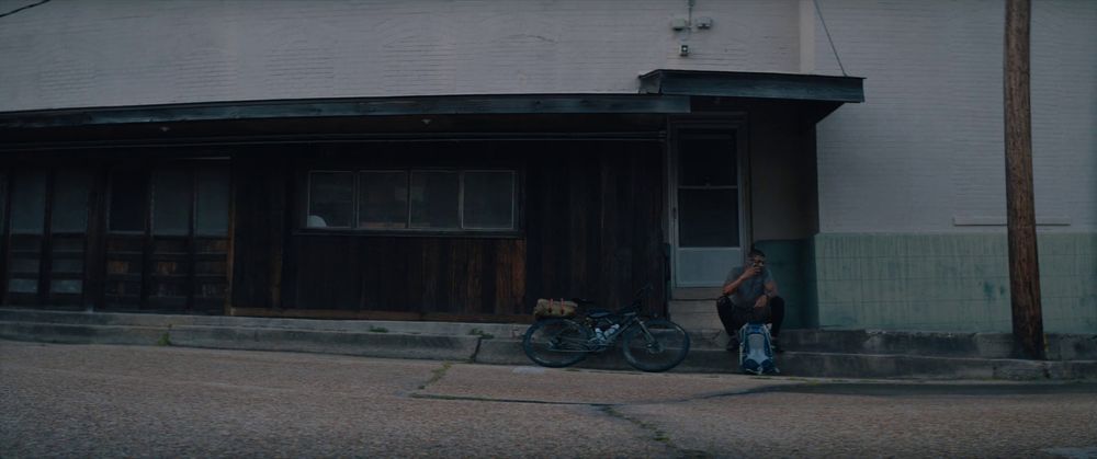 a rundown building facade, in dark-stained wood, inset in a wall of off-white painted empty. a black man sits beneath the overhanging roof that juts out above the white door, in the shade, his bike beside him and his backpack between his sprawled legs. he does not look like the call he just made has left him any more comfortable than the brief respite he's taken from the sun.