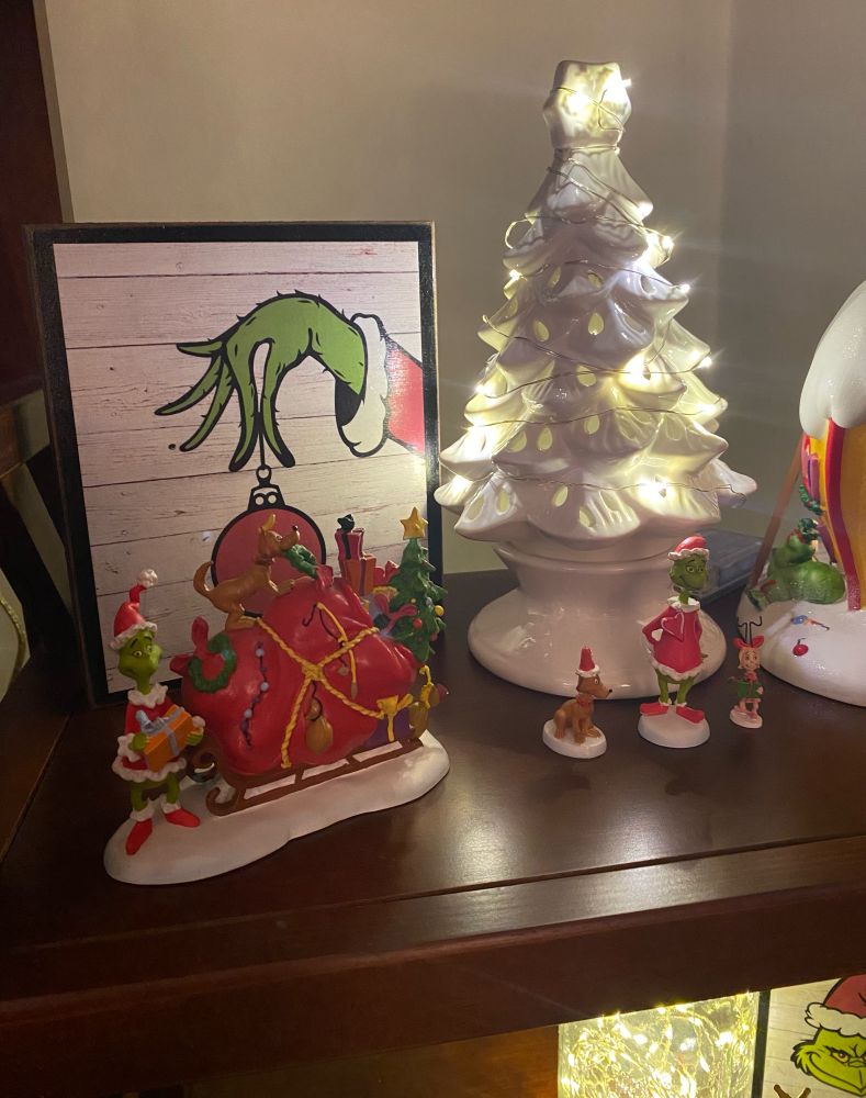A display with a wooden plaque showing the Grinch’s hand holding an ornament, a white ceramic tree with fairy lights, mini figurines of the Grinch, Max and Cindy Lou Who, and a figuring showing the Grinch next to his sleigh that is overstuffed stolen gifts and decorations, with his dog Max perched on top. 