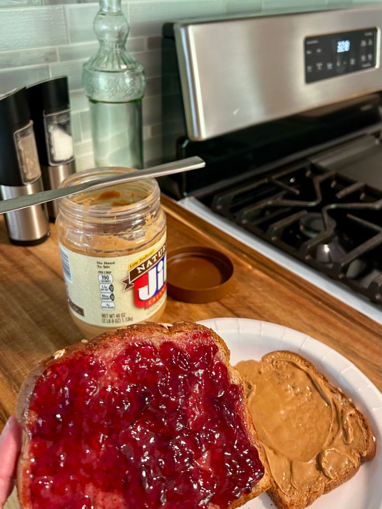 A grape jelly and peanut butter sandwich . Also a jar of peanut butter , a set of salt & pepper grinders , and a pretty jar of olive oil all on a wooden cutting board .