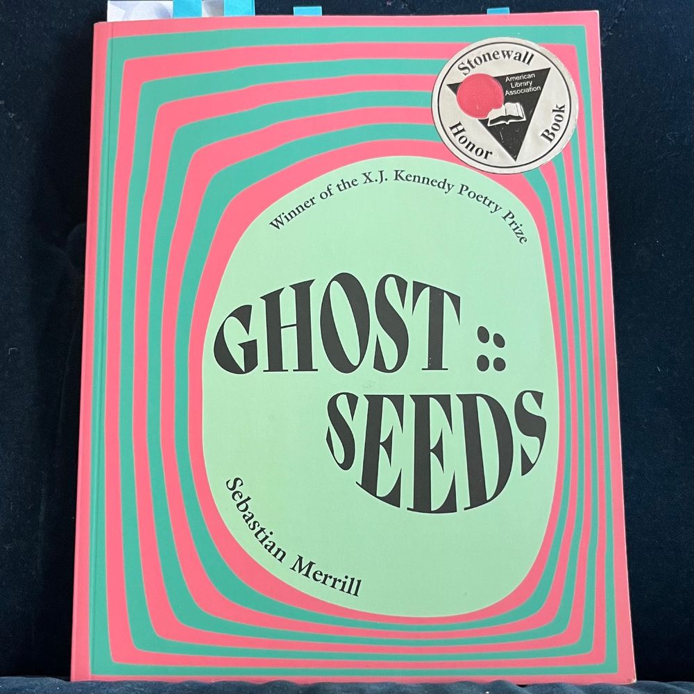 [book sits on blue velvet couch]

All text is in curving black on a pastel green river-rock-shaped space, slightly to the right & downward on the cover. The rock is surrounded by concentric bands of alternating pink & turquoise rippling outward to the edges (& becoming more rectangular as they travel outward). 

[small print following the upper rounded edge of the rock:]
Winner of the X.J. Kennedy Poetry Prize

[huge, centered & growing big then small then big then small to echo/fit the rock edges:] 
GHOST ::
      SEEDS

[medium size, in the lower left round corner of the rock:]
Sebastian Merrill

A circular silver seal in the upper right corner (with a black triangle, an open book inside, & a large pink dot over the left edge of the book & triangle corner) reads:
Stonewall Honor Book
American Library Association