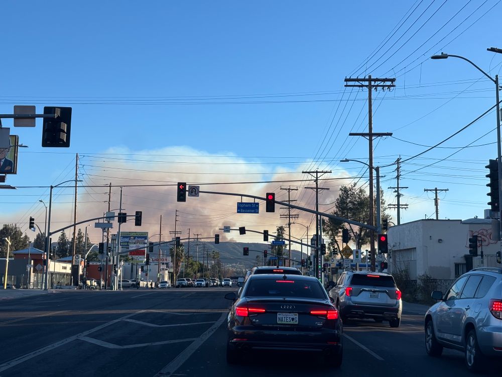 View of a street with cars and a plume of smoke rising over mountains in the background. 