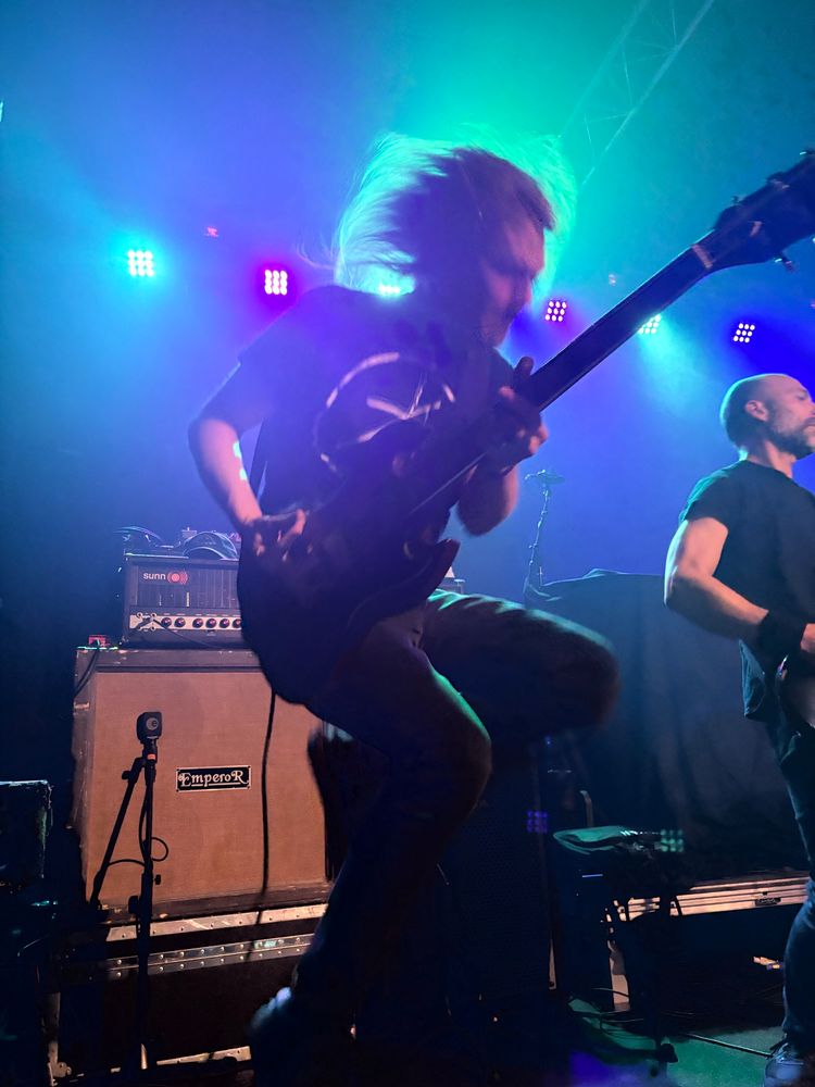 A man playing guitar, blurry with motion, hair flying as he jumps. There are amps behind him, and a stoic bass player beside him.