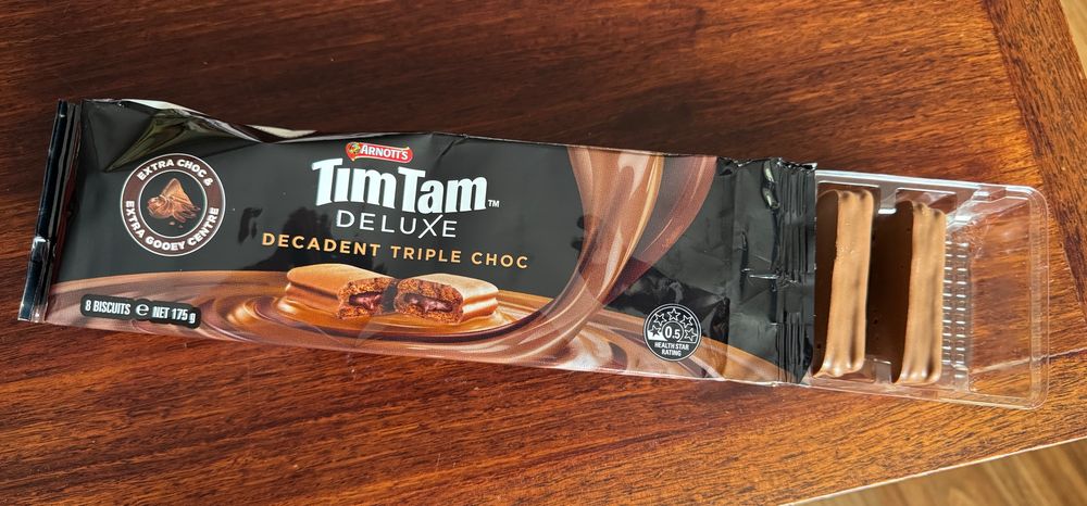 Packet of Arnotts TimTam Deluxe Decadent Triple Choc biscuits. Badge says “Extra Choc, Extra Gooey Centre”. Biscuits are visible protruding from wrapping.