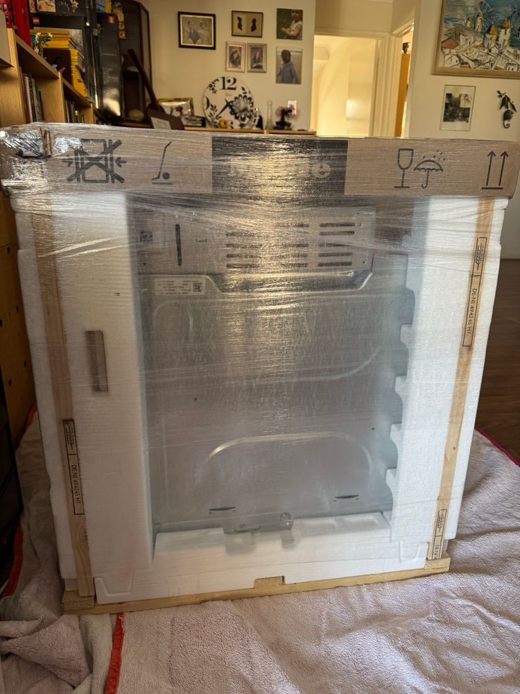 View of oven on a small pallet wrapped in styrofoam etc. Cardboard has Miele and various hazard symbols printed on it.