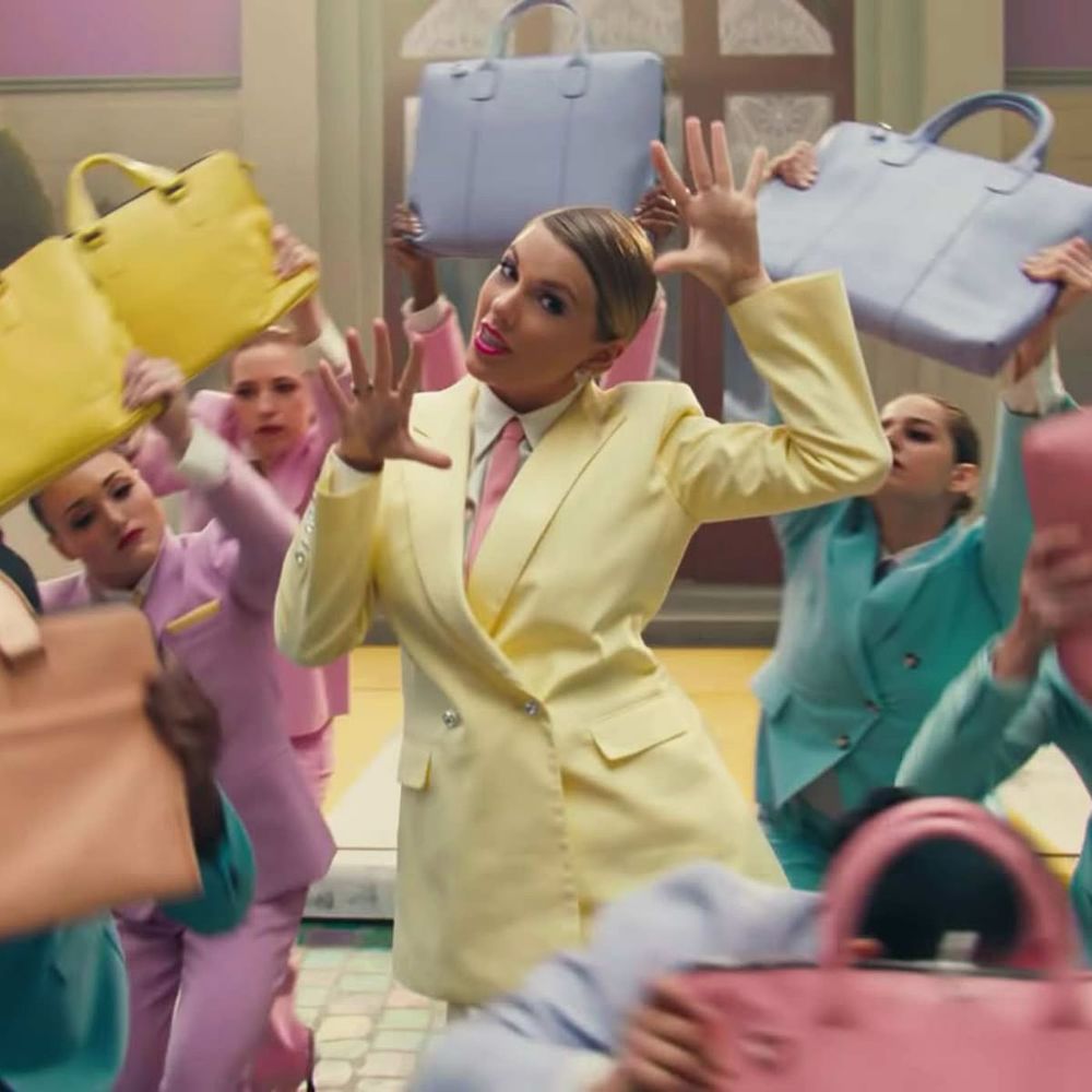A still from Taylor Swift’s “ME!” music video; Taylor stands in a yellow suit surrounded by dancers in pink and teal suits. 