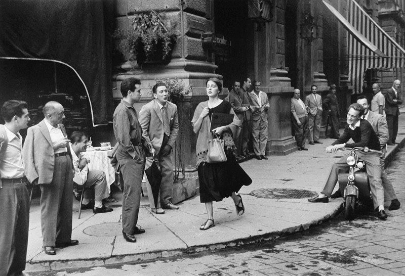 Monochrome photograph featuring a woman walking down a street as men of diferent ages look at her or seem to want her attention, including blociing her path 