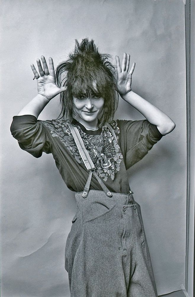 Monochrome photograph featuring a standing white woman holding her hand up to her heaf with palms facing firwards, smiling, with dark spikey hair,  wearing a patterned top and trousers