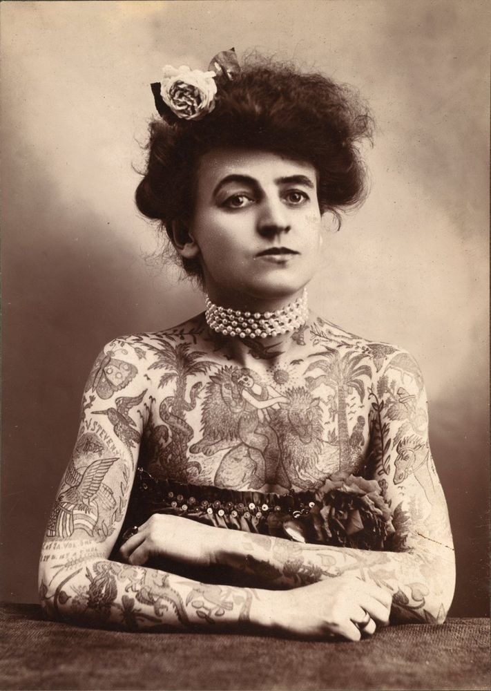 Monochrome photograph featuring a sitting white woman, upper half facing forwards, with chest and shoulders partly bare to reveal many tattoos, her arms are slightly folded, she wears a pearl necklace and has a flower in her dark hair