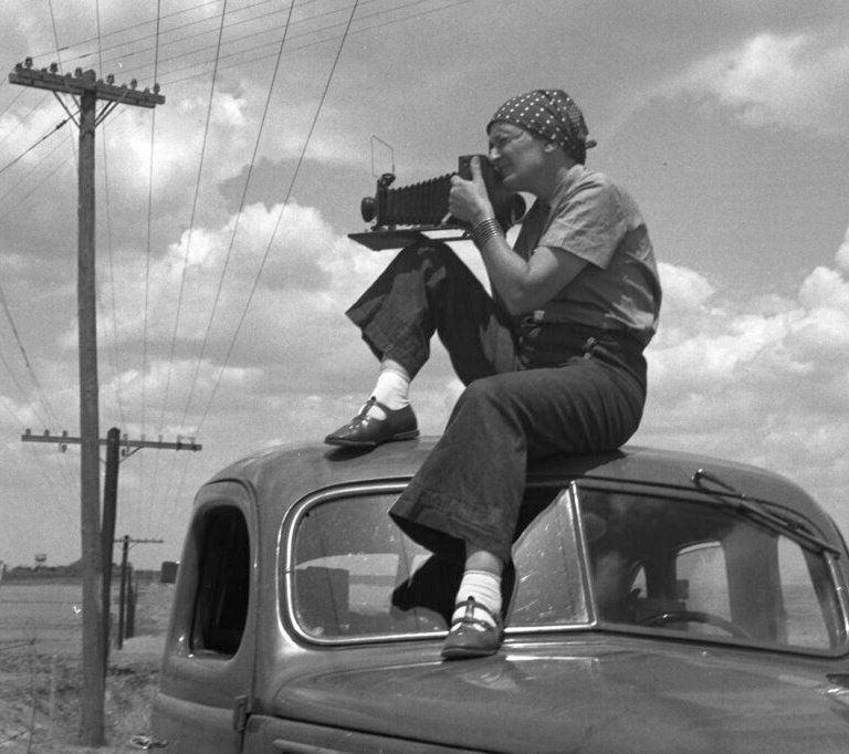 Monochrome photograph featuring a white woman holding a large camera to her face while sitting on the roof of a 1930s style car