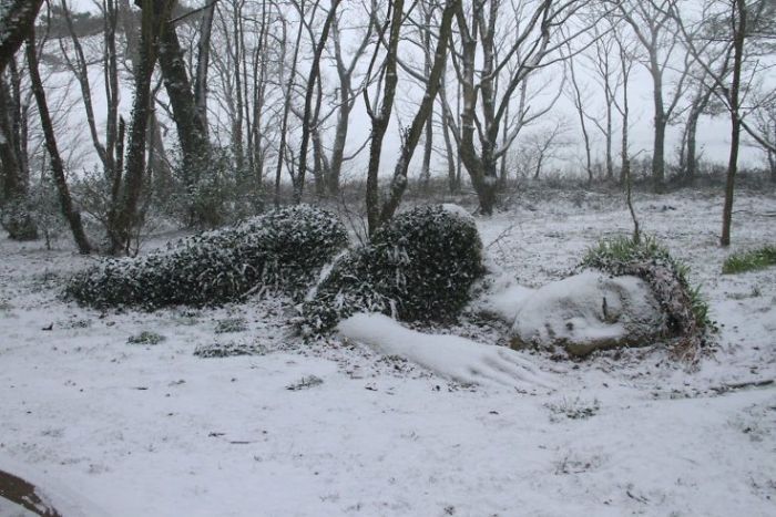 Photo of a sculpture in a rural location created to look like a woman lying on the ground, as if sleeping on her side with only the right half of her body exposed, the ground and sculpture is covered in frost and snow and a row of bare tees is to the background 