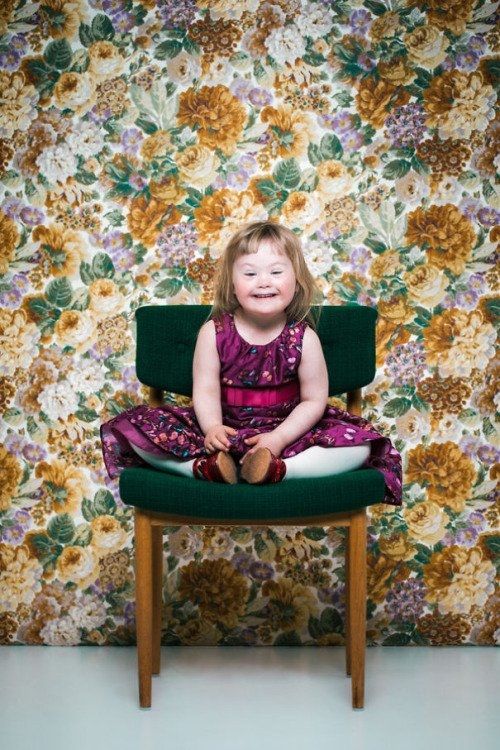 Photo of a very young white girl sitting on a chair in front of floral wallpaper  with legs up facing forwards, she wears a pink patterned dress, has blonde hair and is smiling