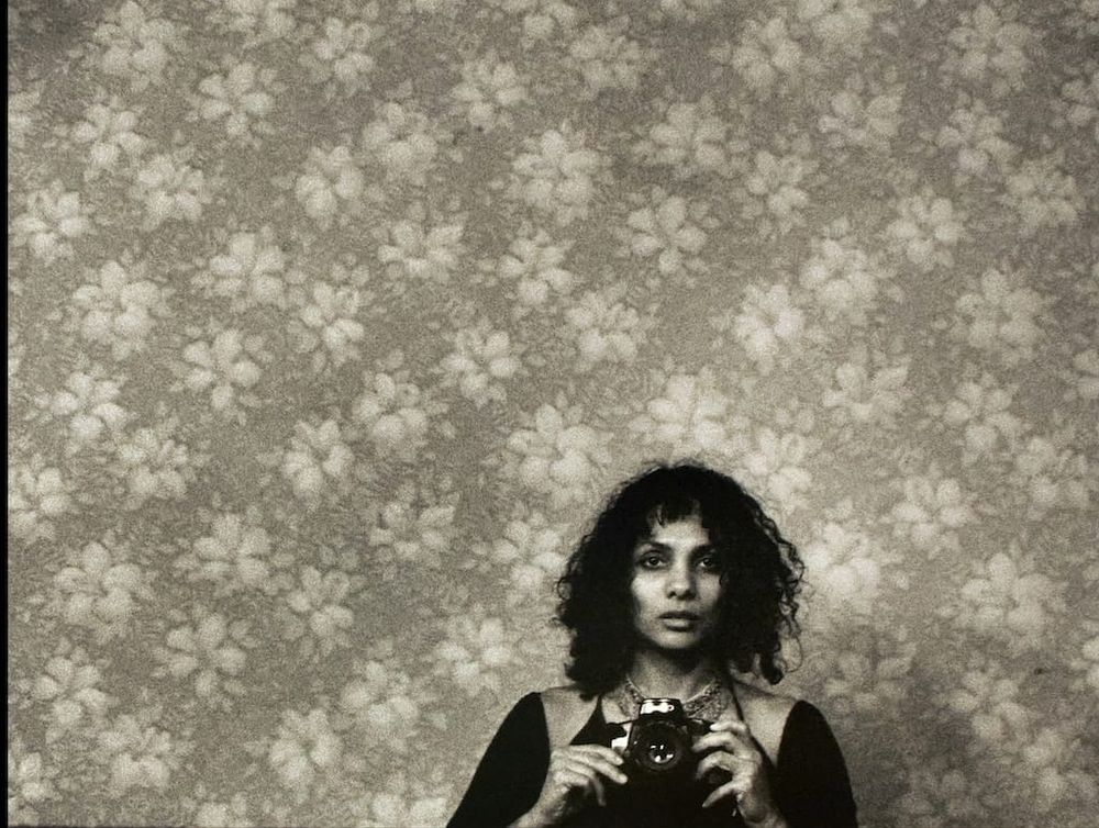 Monochrome photograph of the head and shoulders of a Black woman with long waved hair facing forwards holding a camera, the subject stands against a wall with floral wallper