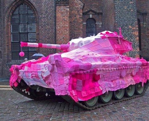 Colour photograph of a tank pointing left on a street in front if a brick building, the tank is covered in a blanket of crocheted or knitted yarn in various shades of pink with a fluffy pink pom pom handing off the gun barrel 