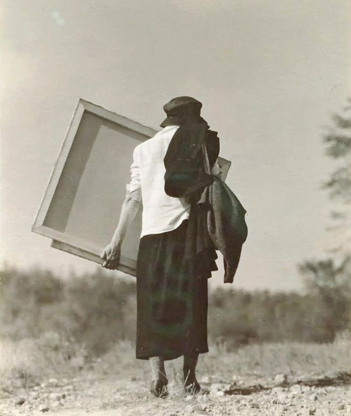 Monochrome photograph featuring a white woman in a long dark skirt, white shirt and black hat, walking with back to the viewer carrying a large canvas on rocky ground with shrubs