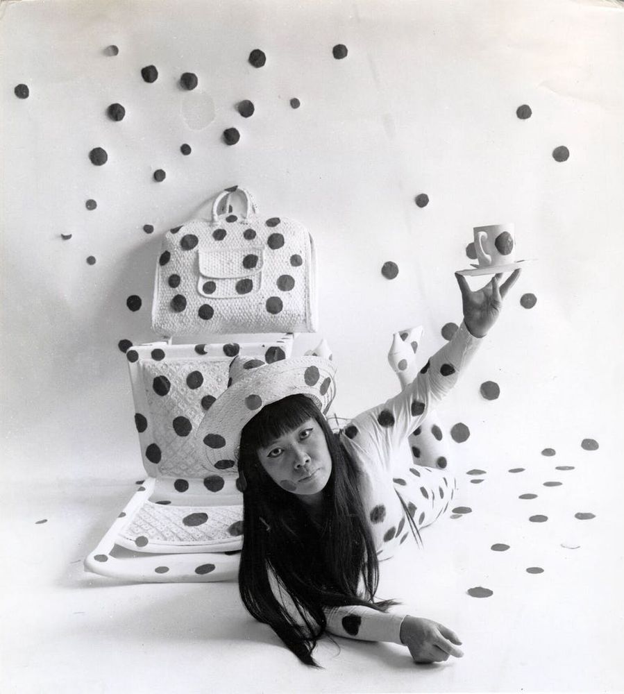 Monochrome photograph featuring a Japanese woman lying down facing forwards with one arm in the air, dressed in white with black dots, next to white objects covered in a similar way as well as dots on the white floor and wall