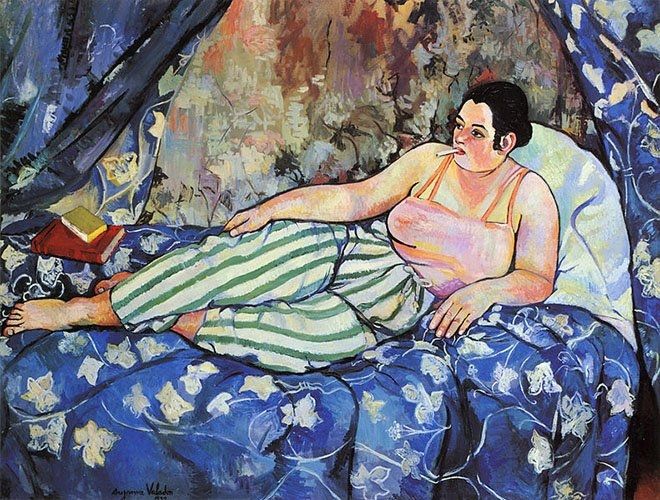 Painting of a woman lounging on a bed with blue covers, she wears a pink vest top and striped pants, she is smoking and a pile of books is near her bard feet