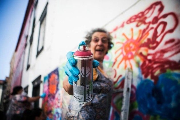 Photo of a street with buildings the right and a board covered in spray painted designs, in the foreground is an elderly white woman looking outwards holds a spray paint container